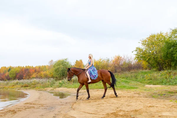 En flicka i en aftonklänning hoppar på en häst på stranden av en damm — Stockfoto