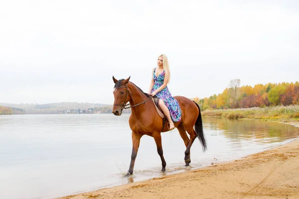 A girl in an evening dress jumps on a horse on the shore of a pond — Stock Photo, Image