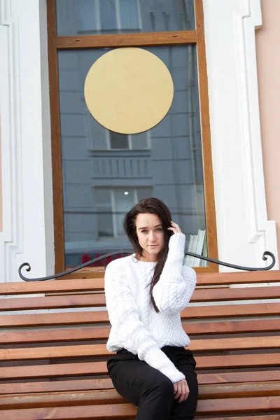 Young girl in a white sweater sits on a bench on a building background — Stock Photo, Image