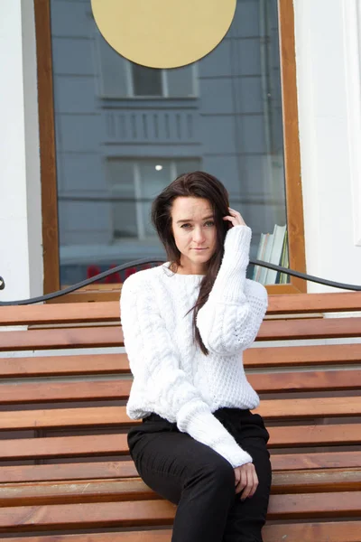 Chica joven en un suéter blanco se sienta en un banco sobre un fondo del edificio — Foto de Stock