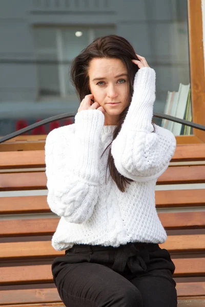 Chica joven en un suéter blanco se sienta en un banco sobre un fondo del edificio — Foto de Stock