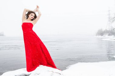 Genç, uzun kırmızı elbiseli kız karlı bir nehir kıyısında duruyor. Kar yağışıweather condition.