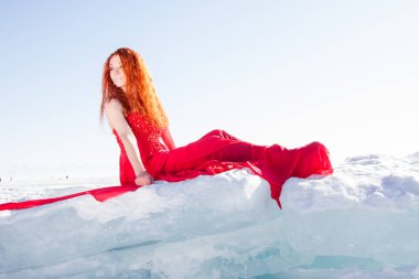 Kırmızı elbiseli kızıl saçlı kız karlı bir buz kütlesinin üzerinde oturuyor..