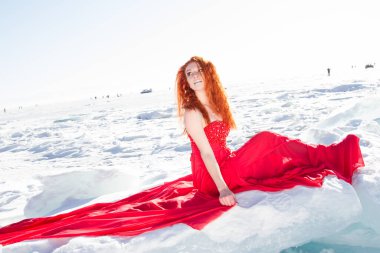 Kırmızı elbiseli kızıl saçlı kız karlı bir buz kütlesinin üzerinde oturuyor..