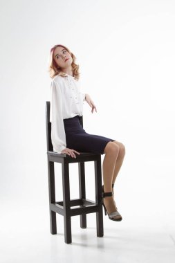 Beyaz kazaklı ve siyah etekli sarışın yüksek bar taburesinde oturuyor. Beyaz arkaplanda stüdyo portresi.