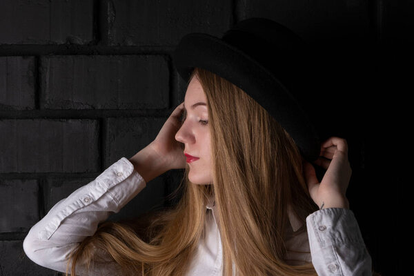 Blonde in a black hat and a white shirt in a dark room.