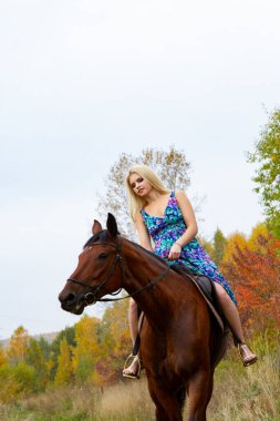 Sonbahar ormanında at süren açık renk bir yaz elbisesi içindeki sarışın.