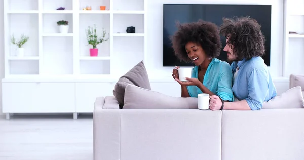多民族のカップルが自宅のソファに座ってコーヒーを飲んでください。 — ストック写真