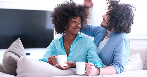 Multietniskt paret sitter på soffan hemma dricker kaffe — Stockfoto
