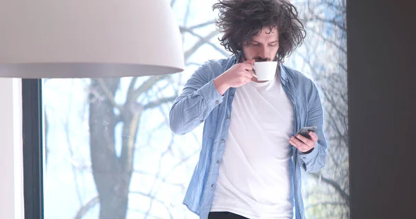 Νεαρός άνδρας, πίνοντας καφέ και χρησιμοποιώντας ένα κινητό τηλέφωνο στο σπίτι — Φωτογραφία Αρχείου