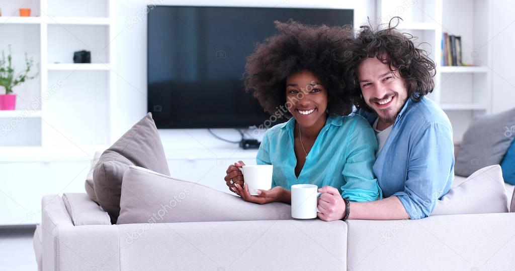multiethnic couple sitting on sofa at home drinking coffe