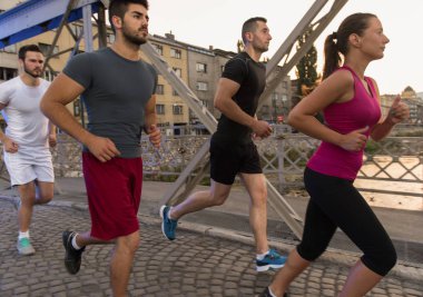 Şehrin güneşli sabahında köprüde koşan bir grup genç sporcu.