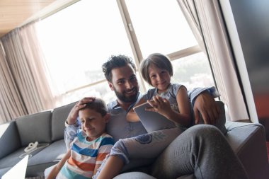 Mutlu bir baba çocuklarla vakit geçirip evdeki koltukta tablet bilgisayar kullanıyor.