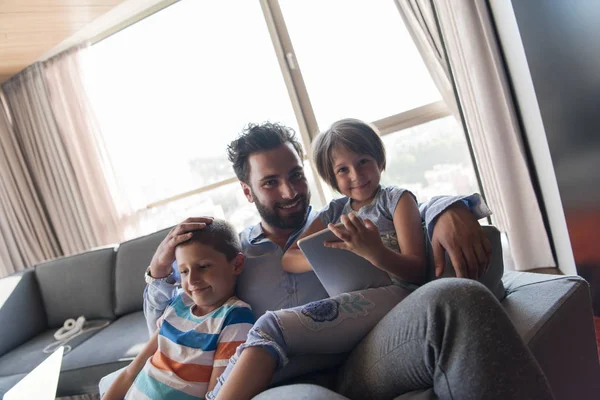 Mutlu bir baba çocuklarla vakit geçirip evdeki koltukta tablet bilgisayar kullanıyor.