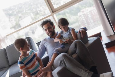 Mutlu bir baba çocuklarla vakit geçirip evdeki koltukta tablet bilgisayar kullanıyor.