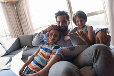 Mutlu bir baba çocuklarla vakit geçirip evdeki koltukta tablet bilgisayar kullanıyor.
