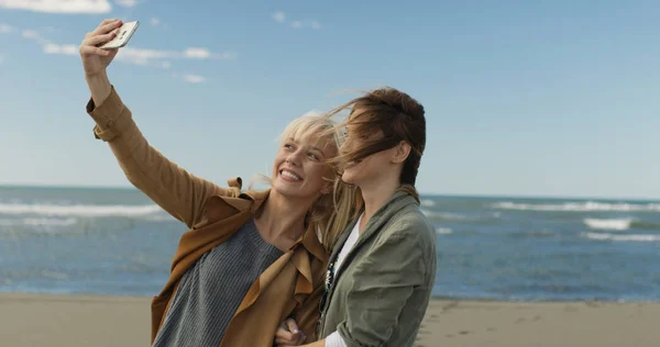 两个微笑的年轻妇女自拍在手机上的秋天在海滩上的一天 — 图库照片