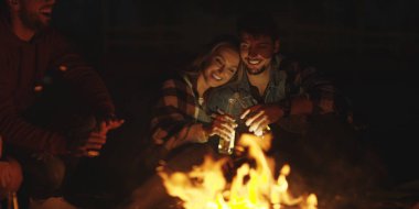 Genç bir çift, arkadaşlarıyla birlikte gece kumsalda oturup bira içiyor.