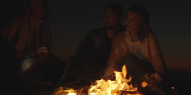 Genç bir çift, arkadaşlarıyla birlikte gece kumsalda oturup bira içiyor.