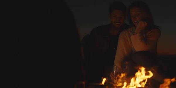 与朋友坐在海滩篝火边喝酒的年轻夫妇 — 图库照片