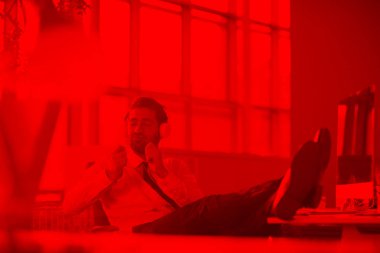 Rahatlamış genç işadamı sabahın erken saatlerinde modern bir ofiste kulaklıkla müzik dinliyor. Arka planda gün doğumu güneşi parlıyor