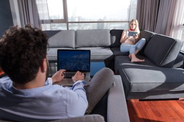 Genç çift kanepede pencerenin yanında tablet ve dizüstü bilgisayar kullanarak rahatlıyor..