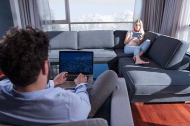 Genç çift kanepede pencerenin yanında tablet ve dizüstü bilgisayar kullanarak rahatlıyor..