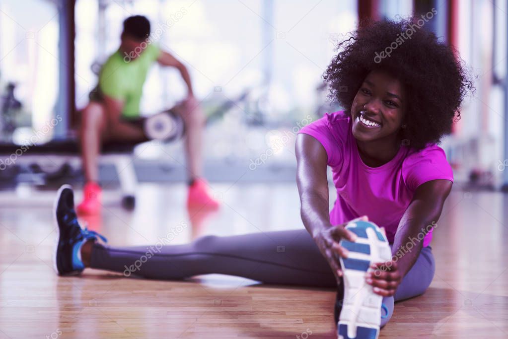 happy young african american woman in a gym stretching and warming up before workout young mab exercising with dumbbells in background