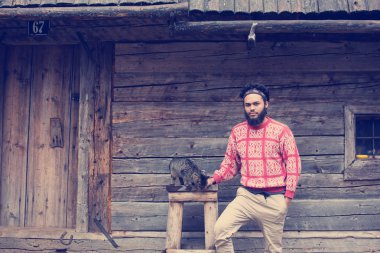 Sakal ve ahşap evin önünde kedi olan genç hipster adam portresi