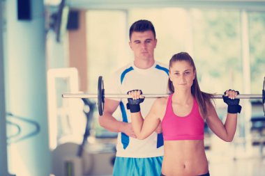 Spor salonunda spor antrenörü olan genç bir kadın ağırlık kaldırıyor.