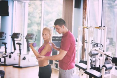 Spor salonunda spor antrenörü olan genç bir kadın ağırlık kaldırıyor.