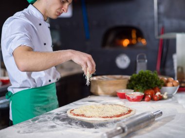 Şef mutfak masasında taze pizza hamurunun üzerine peynir serpiyor.