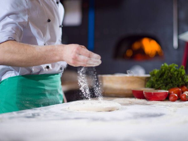 Chef sprinkling flour over fresh pizza dough on kitchen table