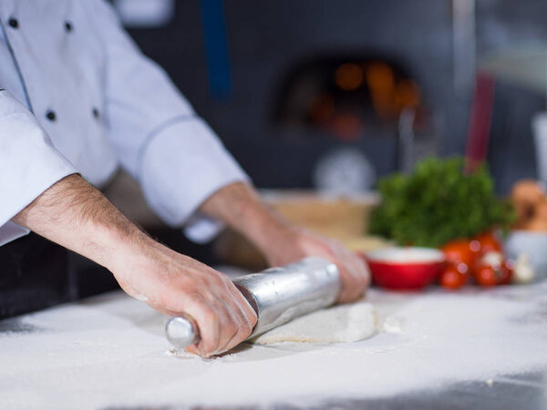 Chef preparing dough for pizza rolling with rolling pin on sprinkled with flour table