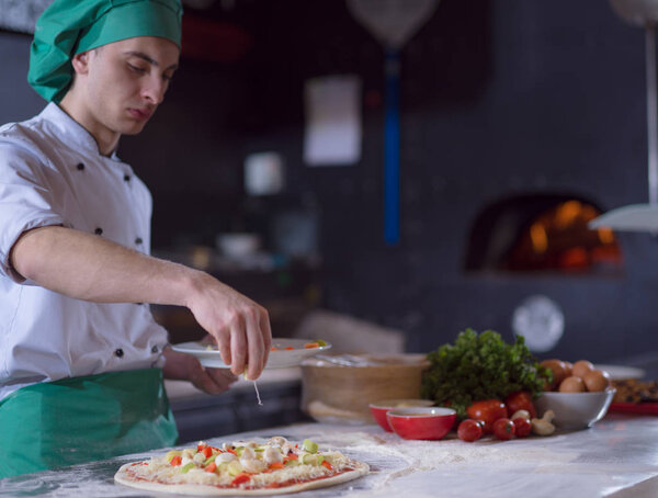 Chef putting fresh vegetables over pizza dough on kitchen table