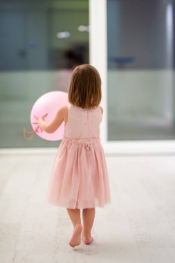 Pembe elbiseli mutlu küçük kız güzel bir akşamda evdeki pencerenin yanında balonlarla oynarken eğleniyor.