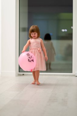 Pembe elbiseli mutlu küçük kız güzel bir akşamda evdeki pencerenin yanında balonlarla oynarken eğleniyor.