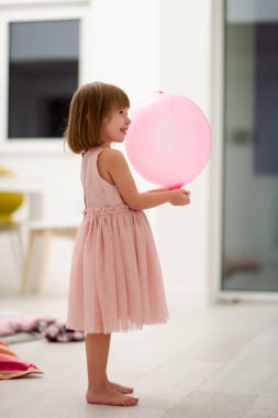 Pembe elbiseli mutlu küçük kız güzel bir akşamda evdeki pencerenin yanında balonlarla oynarken eğleniyor.