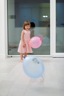 Pembe elbiseli mutlu küçük kız güzel bir akşamda evdeki pencerenin yanında balonlarla oynarken eğleniyor.