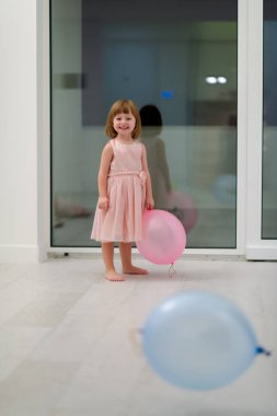 Pembe elbiseli mutlu küçük kız güzel bir akşamda evdeki pencerenin yanında balonlarla oynarken eğleniyor.