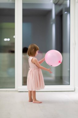 Pembe elbiseli mutlu küçük kız güzel bir akşamda evdeki pencerenin yanında balonlarla oynarken eğleniyor.