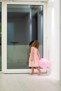 Pembe elbiseli mutlu küçük kız güzel bir akşamda evdeki pencerenin yanında balonlarla oynarken eğleniyor.