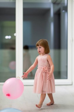 Pembe elbiseli mutlu küçük kız güzel bir akşamda evdeki pencerenin yanında balonlarla oynarken eğleniyor.