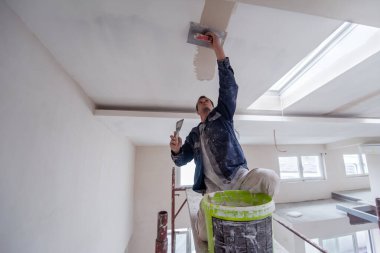 Genç profesyonel inşaat işçisi, alçı taşı tavanını sıva ile sıkarken yeni, modern iki katlı apartman dairesinde darağacını kullanıyor.
