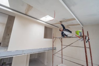 Genç profesyonel inşaat işçisi, alçı taşı tavanını sıva ile sıkarken yeni, modern iki katlı apartman dairesinde darağacını kullanıyor.