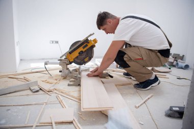 professional worker cutting plank with electrical circular saw during installation a new laminated wooden floor in a unfinished apartment clipart