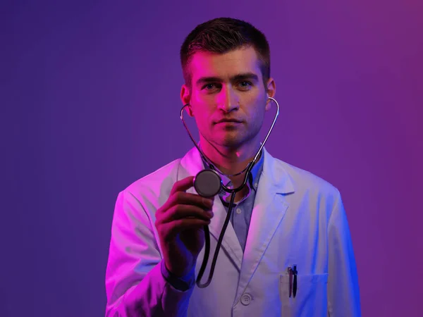 白衣英雄的画像 带着听诊器的快乐微笑的年轻医生站在蓝色和粉色的背景下 Coronavirus Covid 19危险警报 — 图库照片