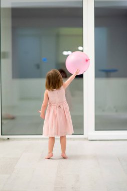 Pembe elbiseli mutlu küçük kız güzel bir akşamda evdeki pencerenin yanında balonlarla oynarken eğleniyor.