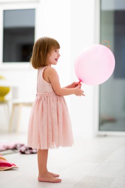 Pembe elbiseli mutlu küçük kız güzel bir akşamda evdeki pencerenin yanında balonlarla oynarken eğleniyor.