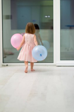 Pembe elbiseli mutlu küçük kız güzel bir akşamda evdeki pencerenin yanında balonlarla oynarken eğleniyor.